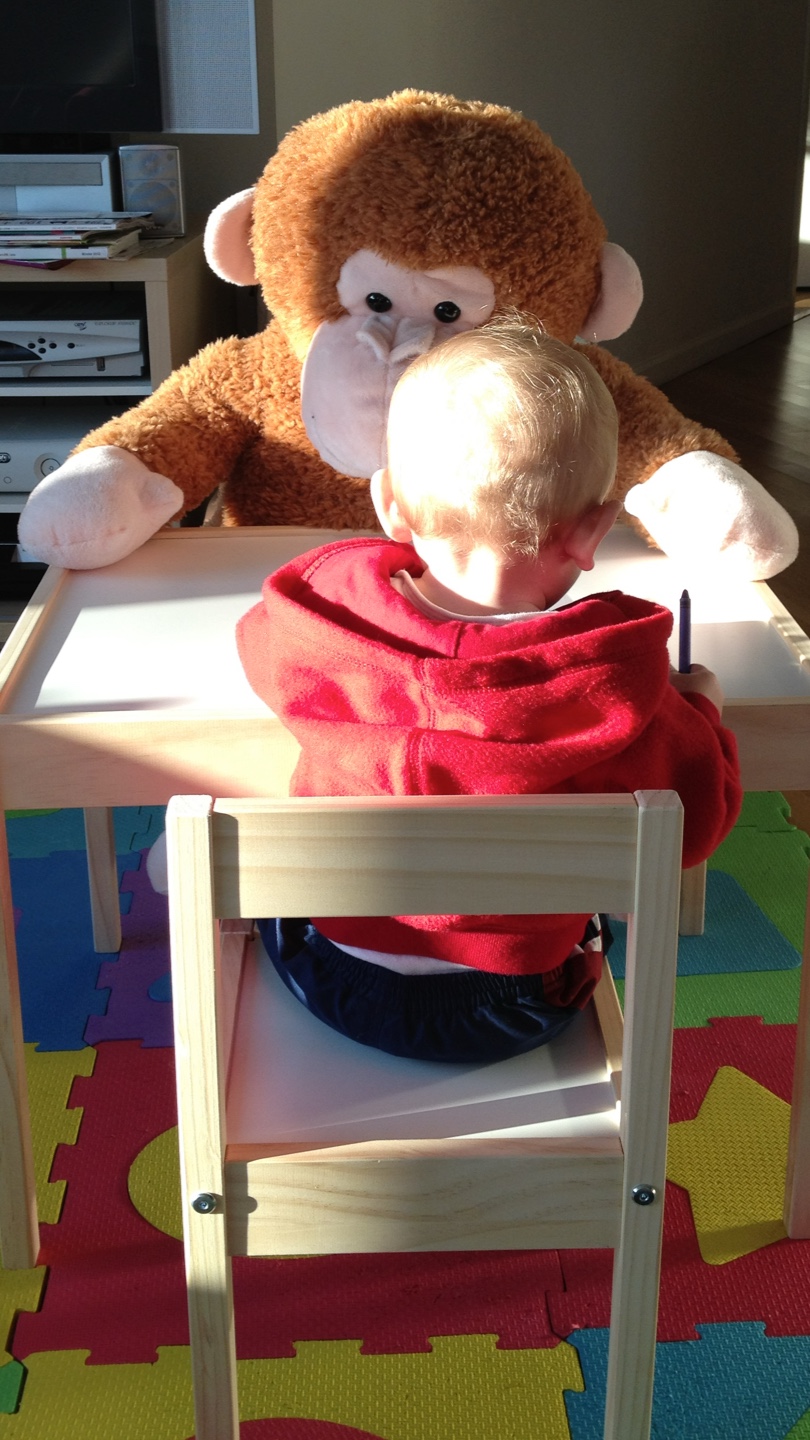 A tofddler boy and a large stuffed monkey at a table drawing.